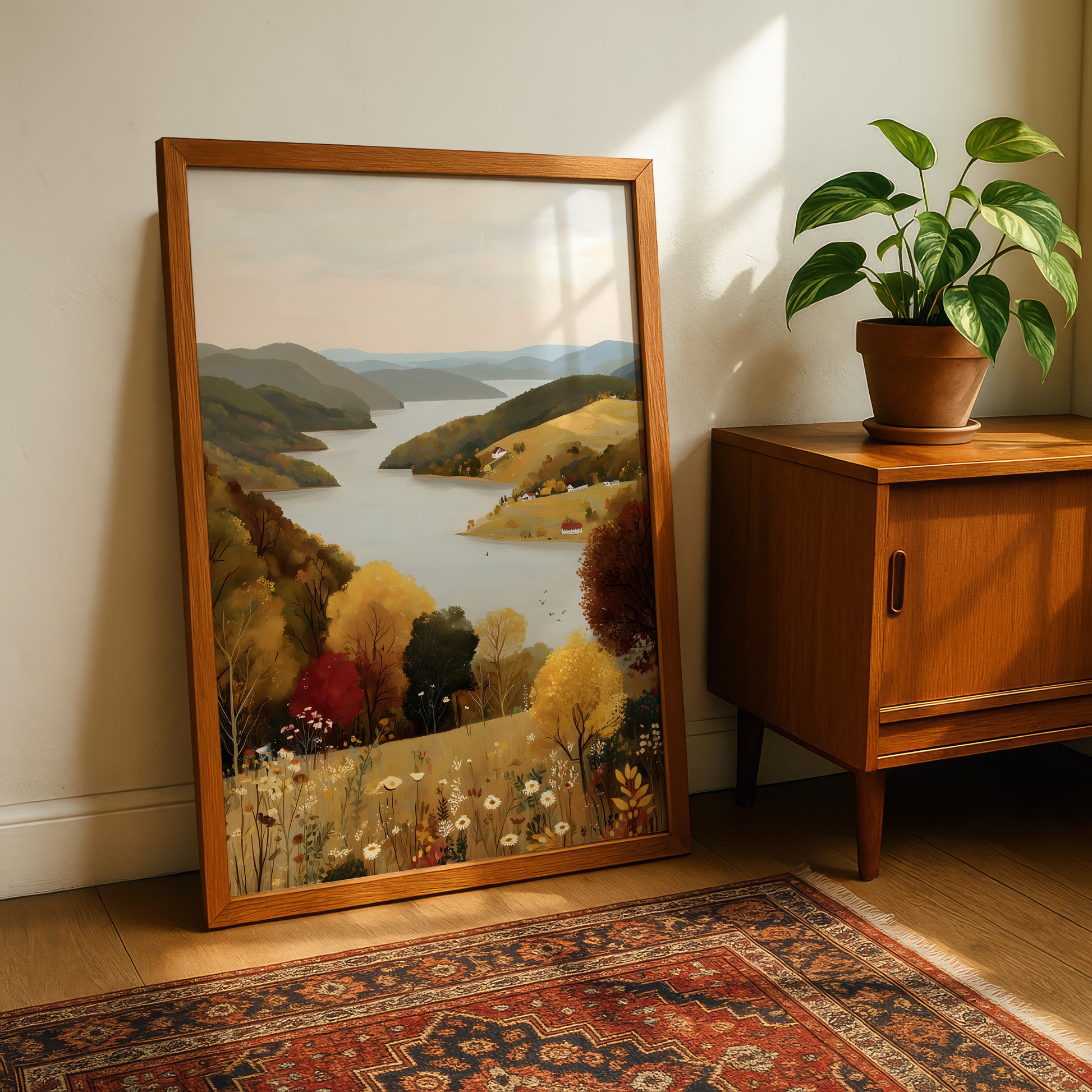a large framed painting of a scenic landscape with a lake, mountains, and trees, placed on a wooden floor next to a potted plant and a wooden cabinet.