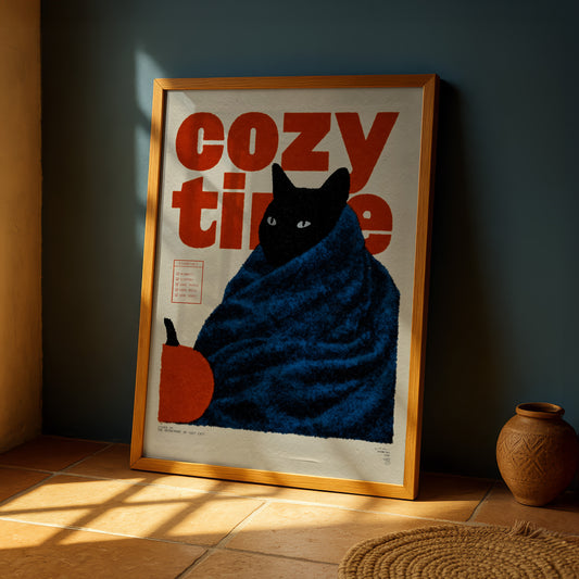 A framed poster with a black cat wrapped in a blue blanket, sitting on a tiled floor next to a vase.