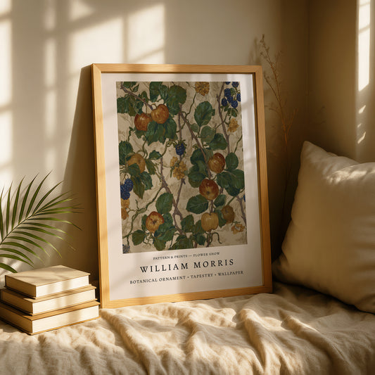 A framed print of a botanical illustration by William Morris, featuring a vibrant floral pattern with various fruits and leaves, is displayed on a bed with a stack of books and a plant nearby.