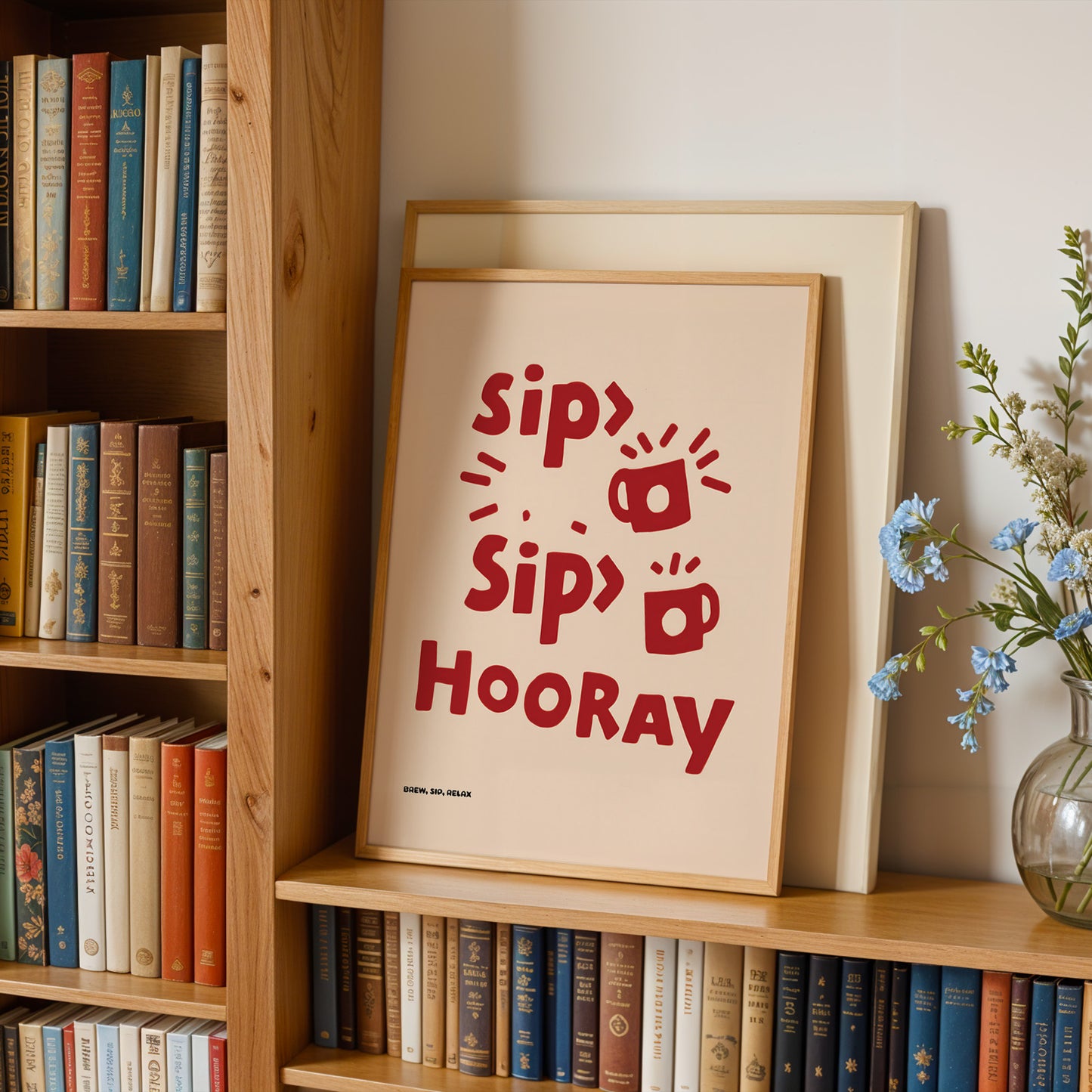 A framed poster with the text "Sip, Sip, Hooray" is displayed on a bookshelf, surrounded by books and a vase of flowers.