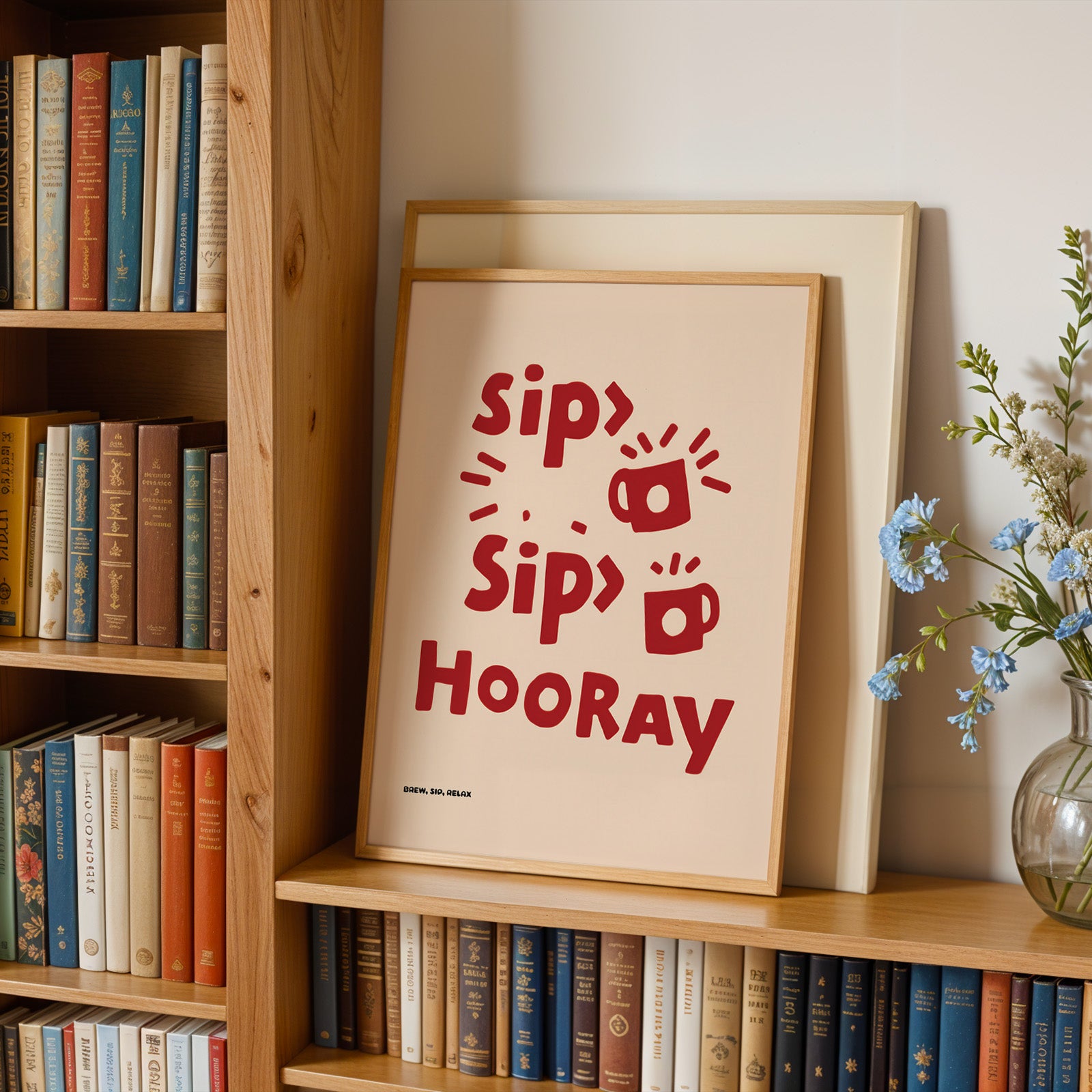 A framed poster with the text "Sip, Sip, Hooray" is displayed on a bookshelf, surrounded by books and a vase of flowers.