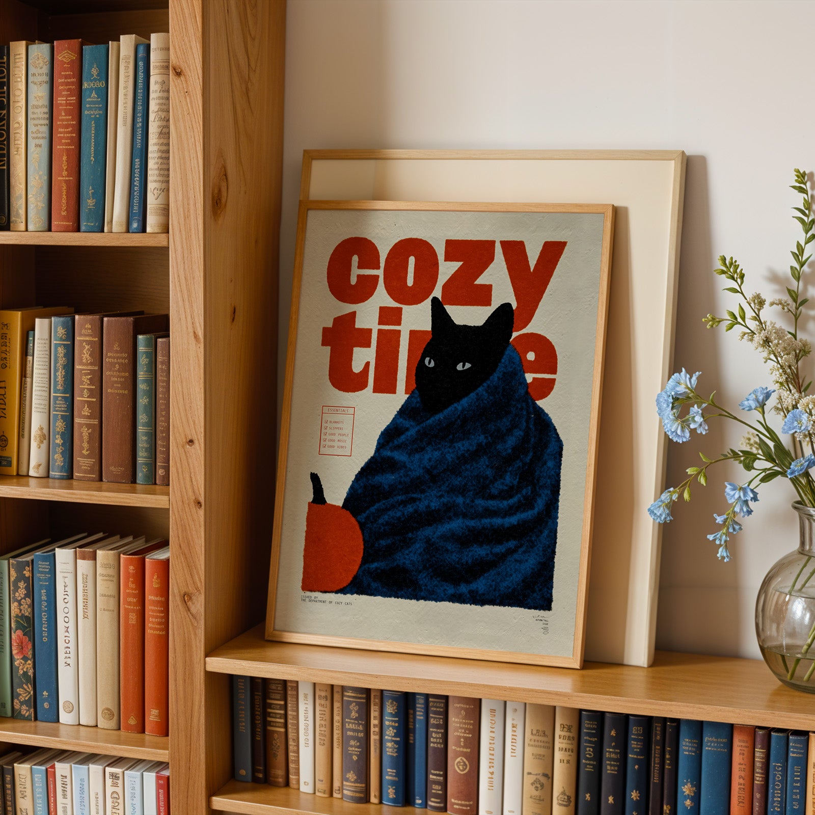 A framed poster of a cat wrapped in a blue blanket, with the text "cozy time" on it, is displayed on a bookshelf next to a vase of flowers.