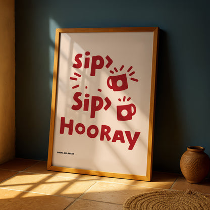 a framed poster with the text "Sip Sip Hooray" in red letters, along with two red coffee cups with rays emanating from them. The poster is leaning against a wall, and there is a woven mat on the floor next to it.