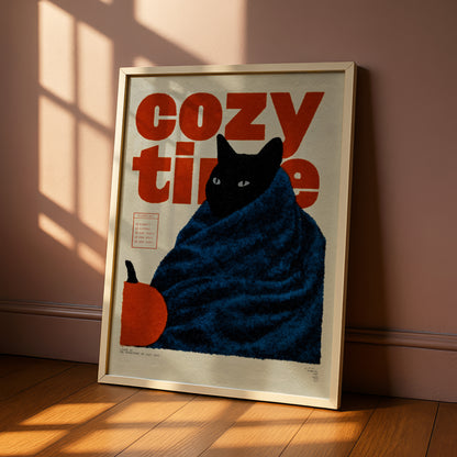 A framed poster with a black cat wrapped in a blue blanket, sitting on a wooden floor and looking out of a window.