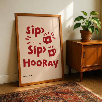 a framed poster with the text "Sip, Sip, Hooray" in red letters, placed on a wooden floor next to a potted plant and a wooden cabinet.