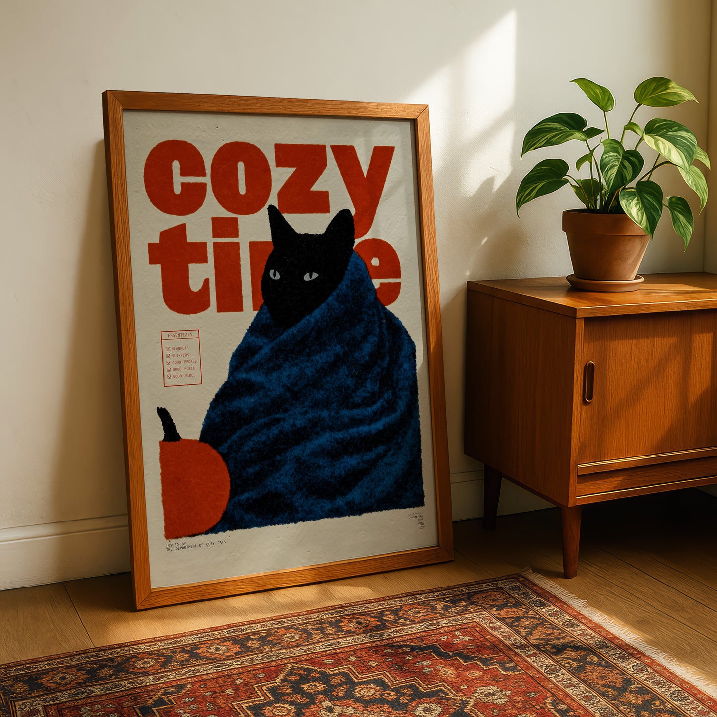 A framed poster of a cat wrapped in a blue blanket, with the text "cozy time" written on it, is displayed on a wooden cabinet next to a potted plant.