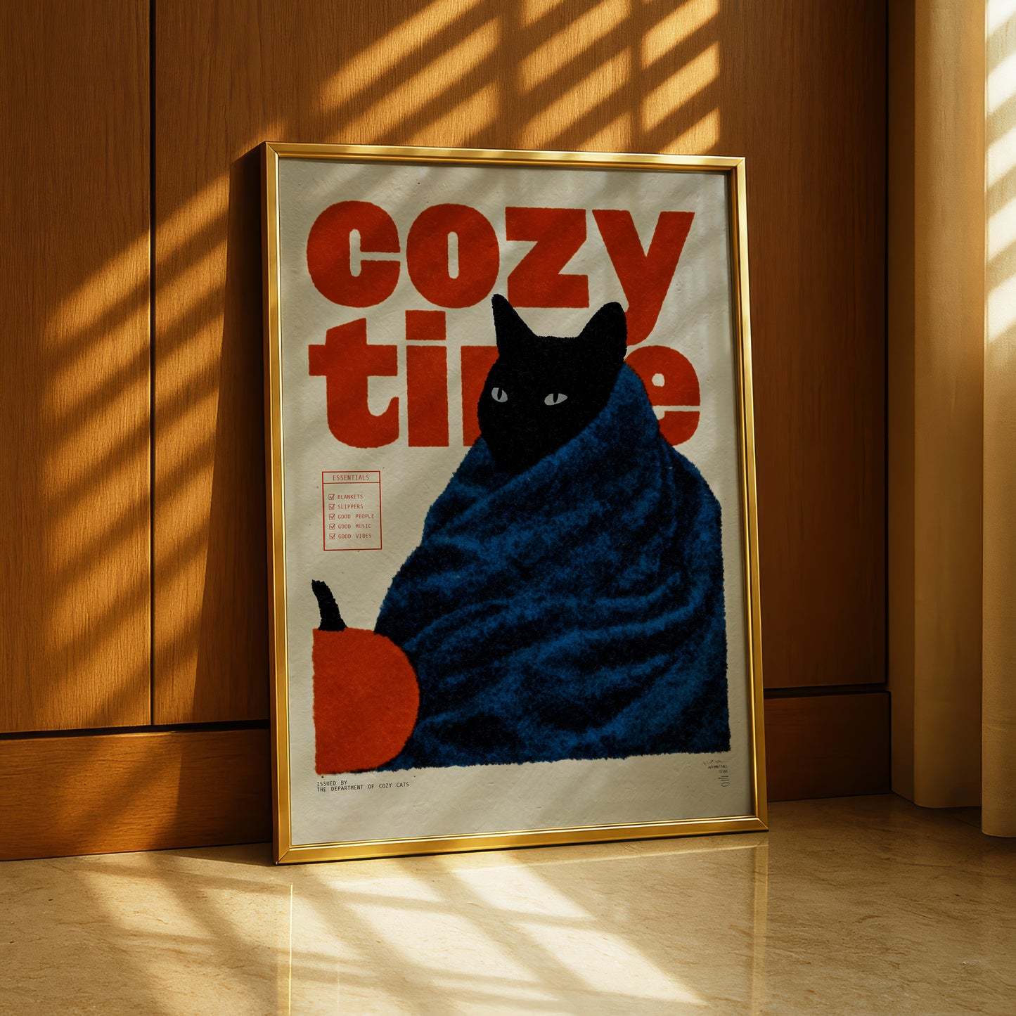A framed poster with a black cat wrapped in a blue blanket, sitting on a wooden floor and holding an orange object.