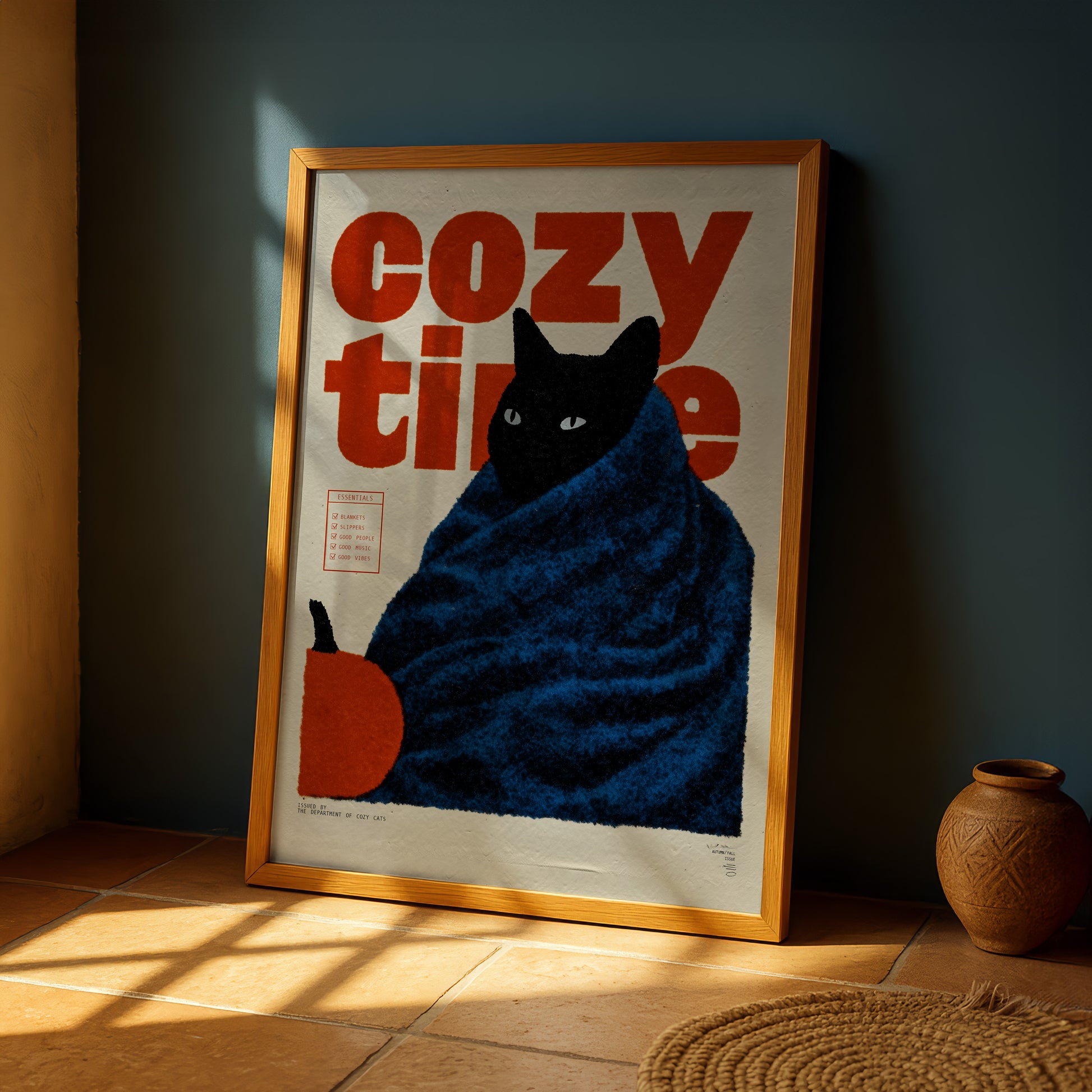 A framed poster with a black cat wrapped in a blue blanket, sitting on a tiled floor next to a vase.