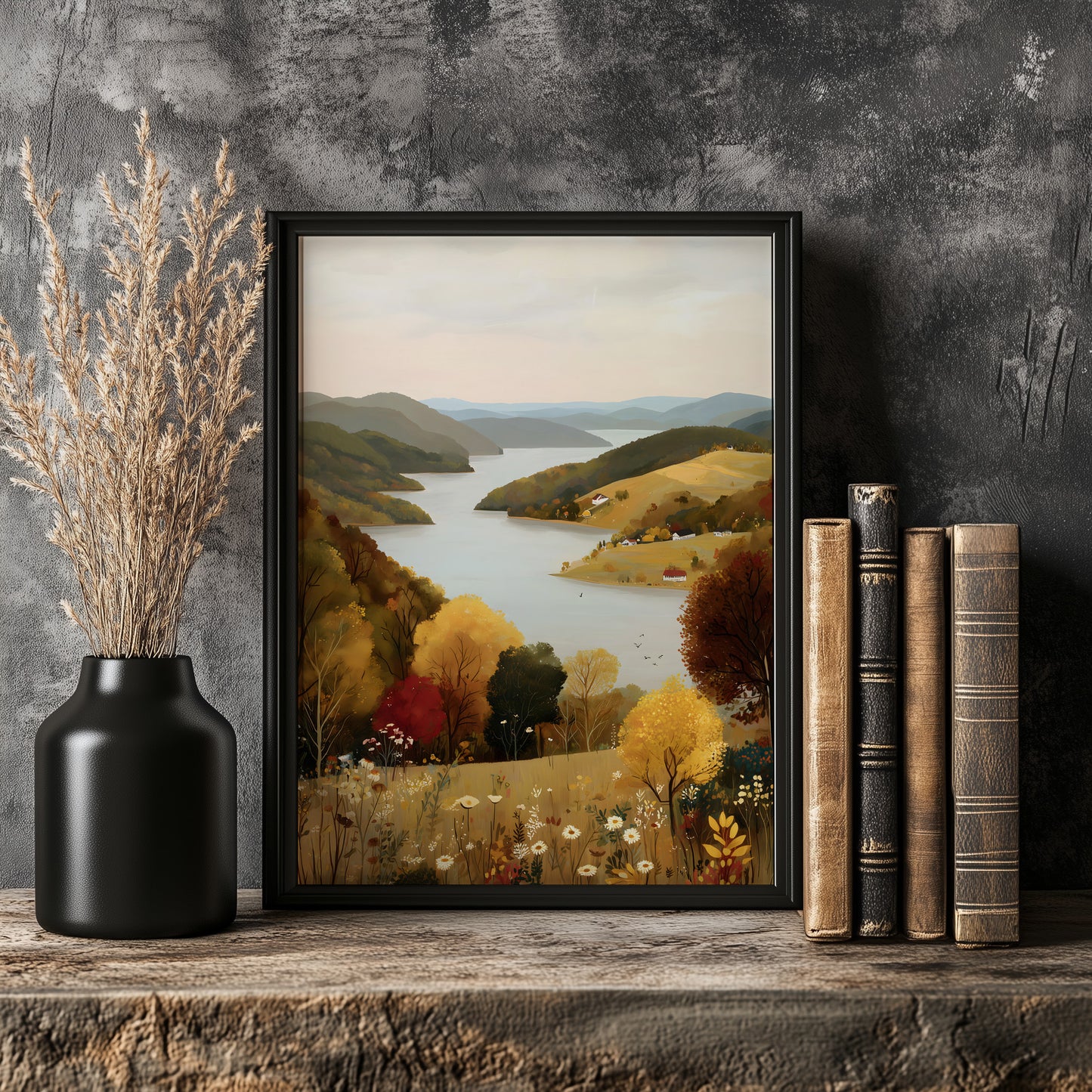 a framed painting of a scenic landscape with a lake and mountains in the background, placed on a wooden shelf next to a vase with dried flowers and a stack of books.