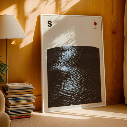 a framed artwork depicting a black and white abstract design, placed on a wooden surface next to a stack of books and a lamp.