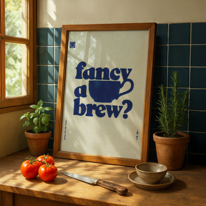 a framed poster with the text " Fancy a brew?" displayed on a wooden table, surrounded by various kitchen items such as a knife, a bowl, and a potted plant.