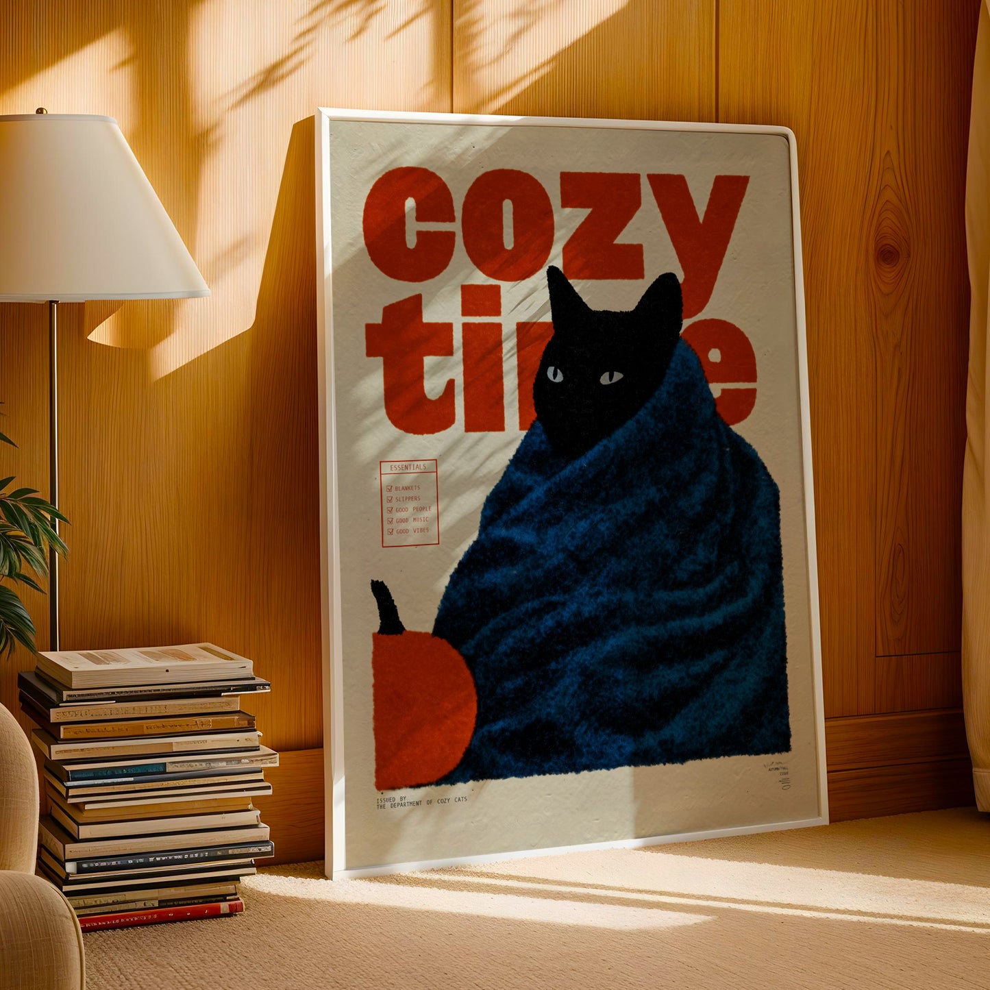 A framed poster of a cat wrapped in a blue blanket, with the text "cozy time" written on it, is displayed on a wall in a room with a lamp and bookshelf.